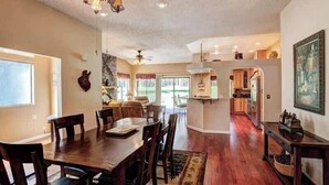 A spacious dining area with a wooden table and chairs, a ceiling fan, and a view of the kitchen and living room through an open door.