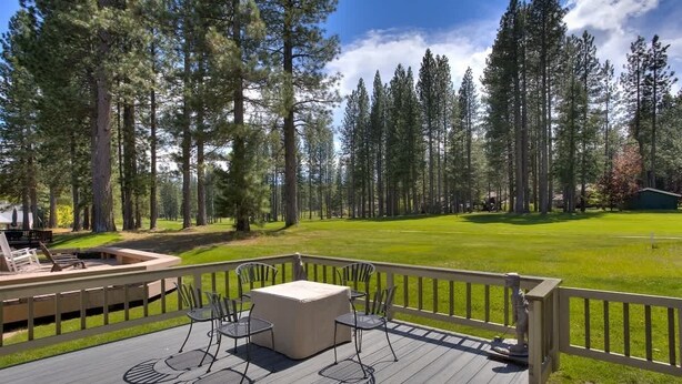 A deck with outdoor furniture, surrounded by a wooden railing, overlooking a well-maintained lawn and tall trees.
