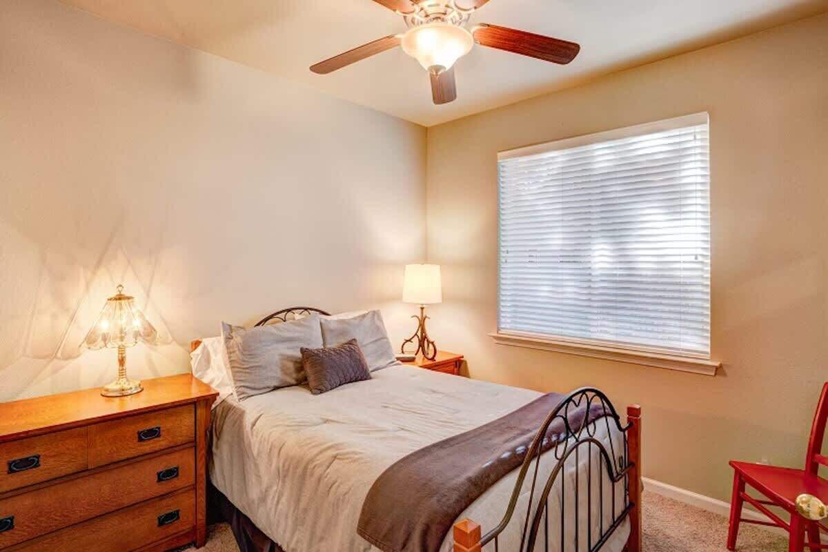 Un dormitorio con cama, mesitas de noche, lámpara, ventilador de techo y ventana con persianas.