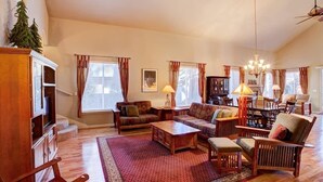 A spacious living room with a high ceiling, wooden furniture, and a large window with curtains. - Mountain Oaks - 35 Oak Court PET! (Blairsden)
