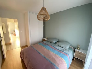 Quarto com cama, mesas de cabeceira, luminária pendente e porta que dá para outro espaço.