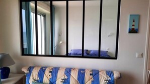 A room with a large mirror, a bed with blue and beige bedding, a small table with a lamp, and a painting of a lighthouse on the wall. - Turpault vue mer port maria quiberon 2* (Quiberon)
