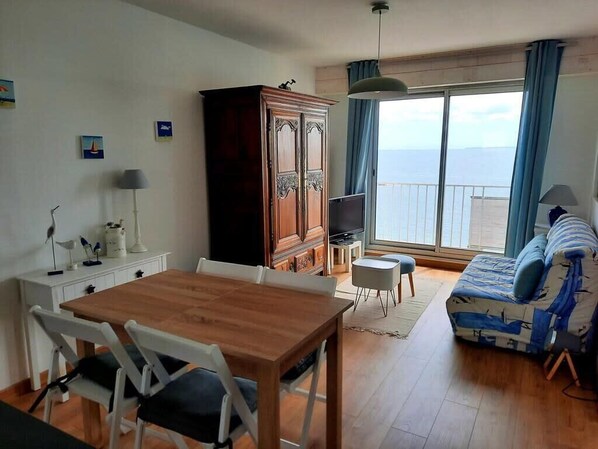 A room with a wooden dining table, white chairs, a sofa, a TV, and a wardrobe.