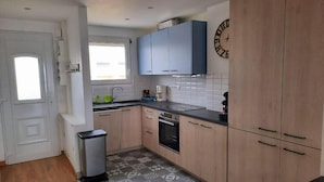 A kitchen with wooden cabinets, a black countertop, a tiled floor, and a clock on the wall. - Turpault vue mer port maria quiberon 2* (Quiberon)