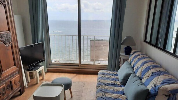 A living room with a sofa, a TV on a stand, a small table, and a view of the sea through large windows. - Turpault vue mer port maria quiberon 2* (Quiberon)