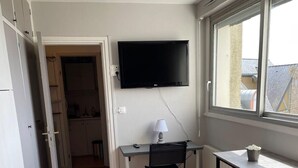 Ein kleiner, gut beleuchteter Raum mit Holzboden, einem Schreibtisch mit Stuhl, einem an der Wand montierten Fernseher und einem Fenster mit Tageslicht.
