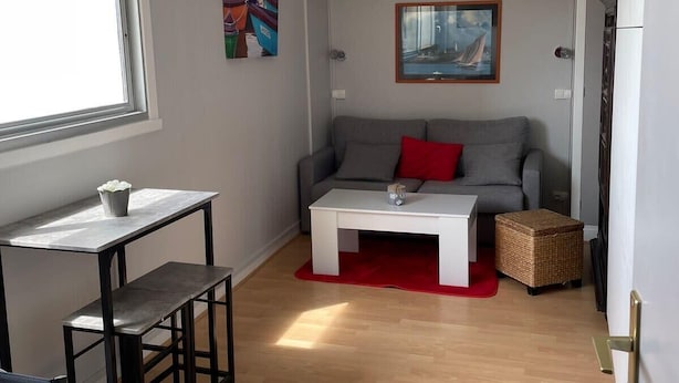 Ein gemütlicher Wohnraum mit grauem Sofa, einem weißen Couchtisch, einem kleinen Tisch mit Pflanze und einem Korbsessel.