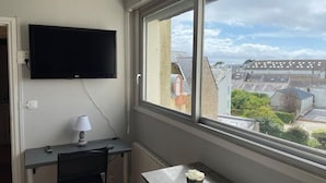 Ein Zimmer mit Schreibtisch, Stuhl, Wandfernseher und Fenster mit Blick auf Gebäude und Bäume.