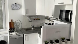 Een moderne keuken met witte kastjes, een ingebouwde magnetron, een spoelbak en een kleine koelkast. Op het aanrecht staan potplanten en erboven hangt een witte hanglamp.