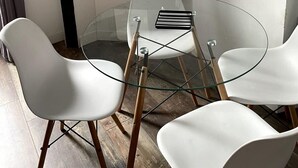 Een kamer met een ronde glazen tafel, witte stoelen, een zwarte console met een spiegel, een klok en een potplant.