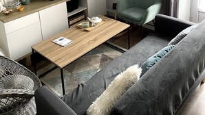 Een moderne woonkamer met een flatscreen televisie, een houten salontafel, een grijze bank en een groene fauteuil. Er staan planken met decoratieve objecten en er hangt een kunstwerk aan de muur.