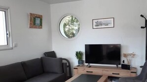 A modern living room with a sectional sofa, a coffee table, and two armchairs. A flat-screen TV is mounted on a wooden stand, and there are two wall-mounted shelves with decorative items. - Maison Tamaris. avec jardin et clos (Quiberon)