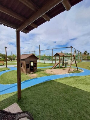 Un parque infantil con columpios y una pequeña estructura de madera, rodeado por una cerca y un camino azul.