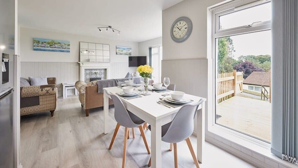 Une cuisine moderne avec des meubles blancs, un coin repas avec une table blanche et des chaises grises, et une grande fenêtre donnant sur l’extérieur.