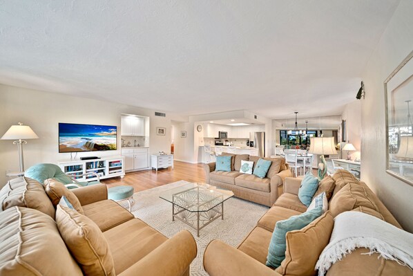 A spacious living room with a large sofa set, a glass coffee table, and a flat-screen TV. A kitchen and dining area are visible in the background.
