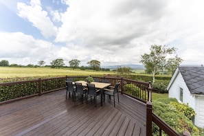 A wooden deck with a table and chairs, overlooking a green landscape and a white building.