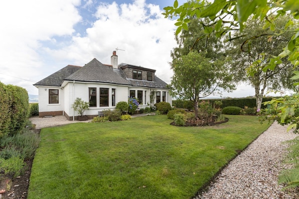 A two-story house with a well-maintained lawn and garden, a gravel driveway, and a view of the sea.