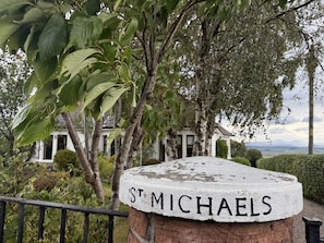 A stone sign with "ST MICHAL'S" inscribed on it, surrounded by greenery and a house in the background.