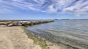 Ein klarer Blick auf einen sandigen Strand mit einem steinernen Wellenbrecher, der sich in ruhiges Wasser erstreckt, unter einem teils bewölkten Himmel.