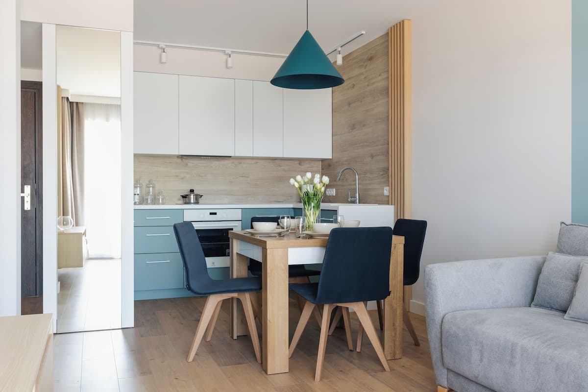 A modern kitchen with white cabinets, a wooden countertop, and a teal pendant light. There is a dining table with two chairs and a vase of flowers.