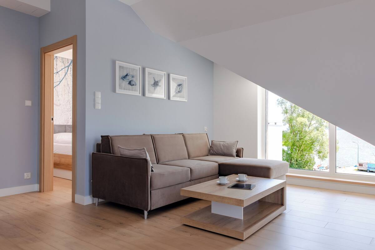 A modern living room with a brown sectional sofa, a wooden coffee table, and a large window offering a view of the outdoors.