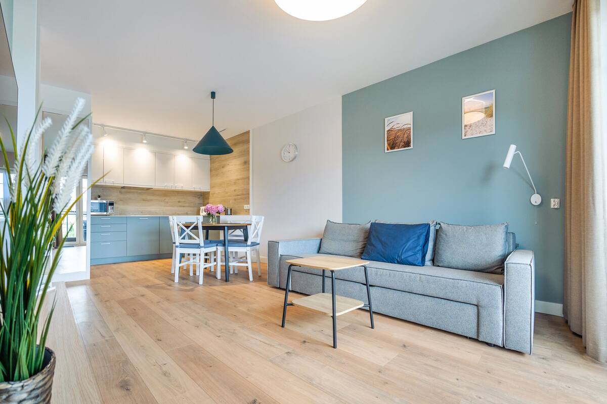A modern living room with a grey sofa, a dining table with chairs, and a kitchen in the background.