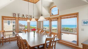 A dining area with a large wooden table and chairs, large windows with a view of the beach, and a 'BEACH' sign on the wall. - Dutch Treat: Oceanfront | Private Pool | Elevator (Corolla)