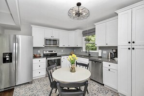 A modern kitchen with white cabinets, stainless steel appliances, a dining table with chairs, and a decorative ceiling light.