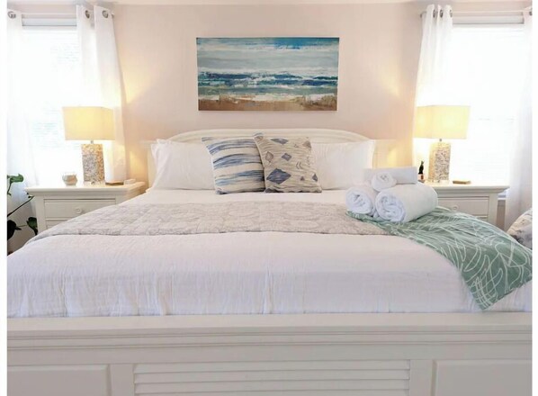 Relax in this serene bedroom with a large bed, adorned with a white comforter and decorative pillows, and a cozy throw blanket.