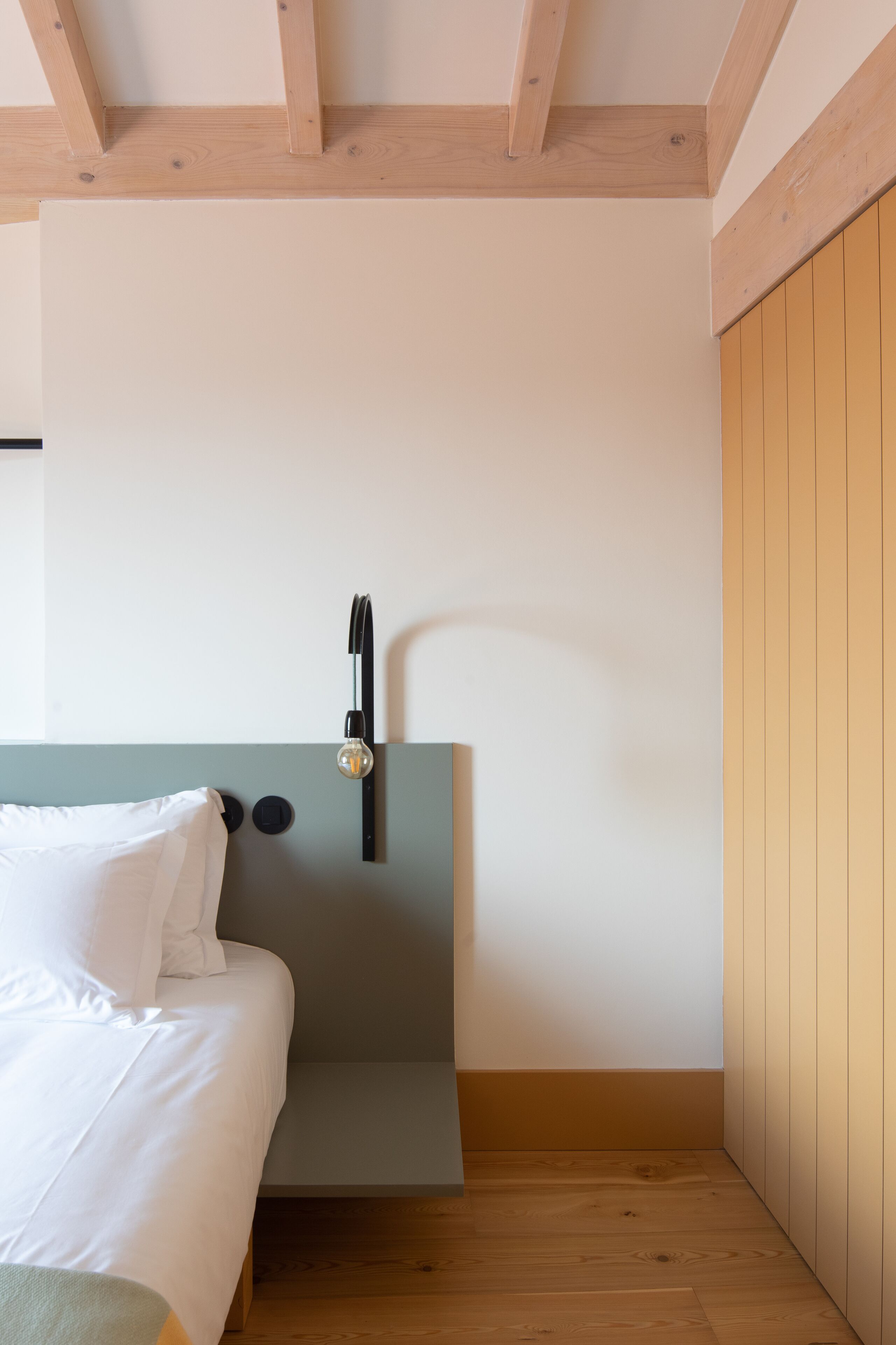 A bedroom with a bed, a bedside table, a wall-mounted light, and wooden flooring.