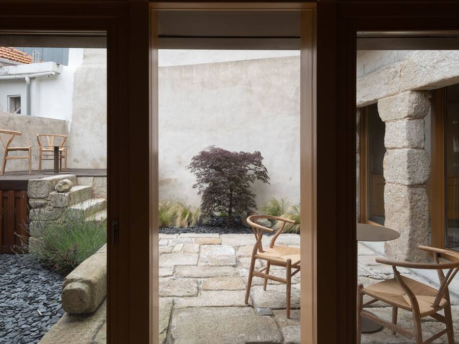 A courtyard with stone flooring, wooden chairs, and a small tree.