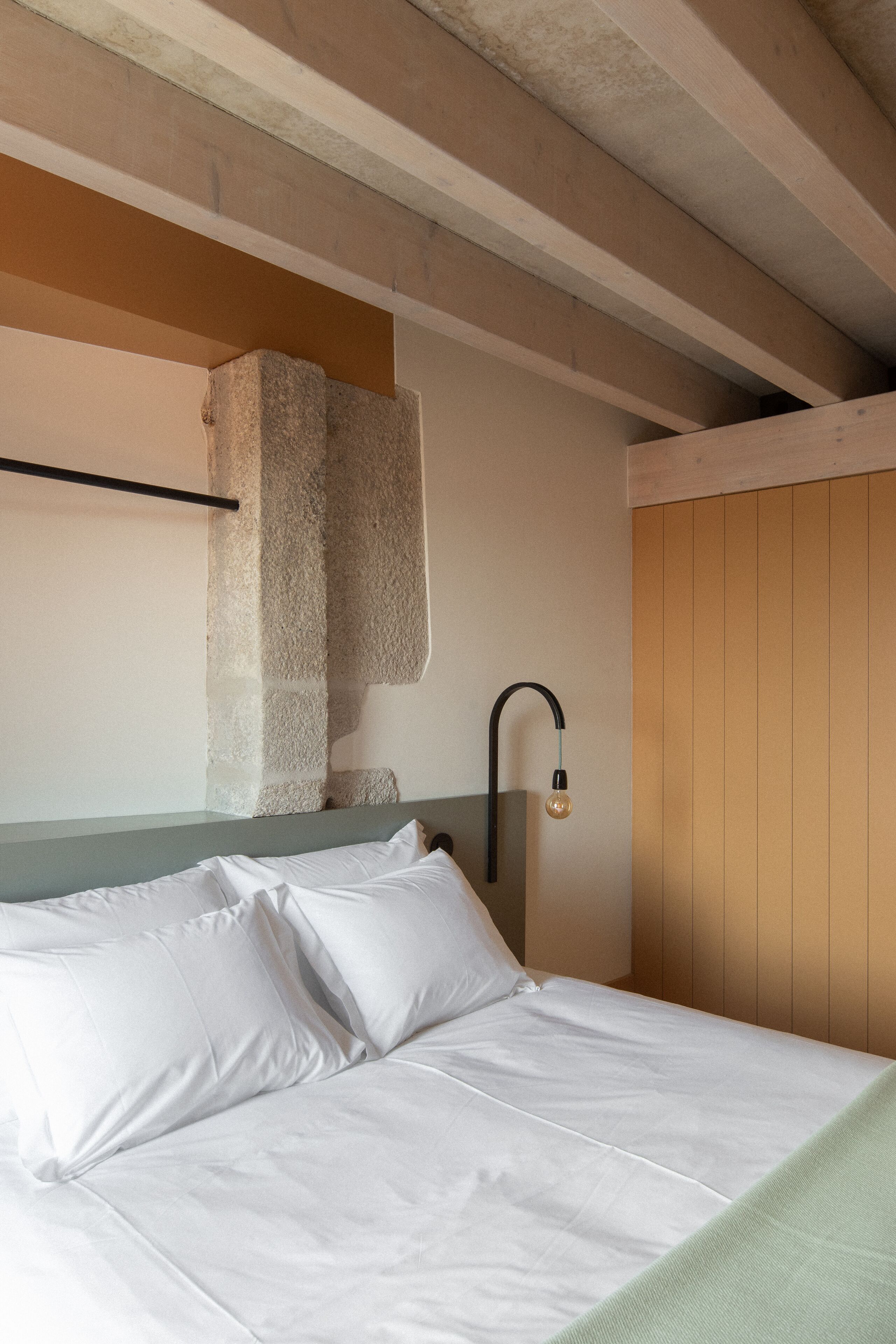 Cama com lençóis brancos e manta verde, cabeceira com detalhe em pedra e luminária fixada na parede.