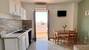 A compact kitchen with a gas stove, white cabinets, and a tiled backsplash. A dining area with a wooden table and chairs is adjacent to the kitchen, featuring a small potted plant. - Sorrento Sunset Home (Meta)
