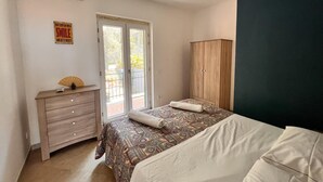 A bedroom with a bed, a wooden wardrobe, a bedside table with a fan, and a window with a view of greenery. - Sorrento Sunset Home (Meta)