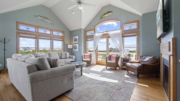 A spacious living room with a grey sofa, a coffee table, wicker chairs, and a fireplace. Large windows offer views of the outdoors. - Soundside Dog Friendly Home w/ Private Pool, Hot Tub, Elevator, and Game Room (Avon)