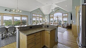 A spacious kitchen with a large island, wooden cabinets, and a view of the outdoors through large windows. - Soundside Dog Friendly Home w/ Private Pool, Hot Tub, Elevator, and Game Room (Avon)