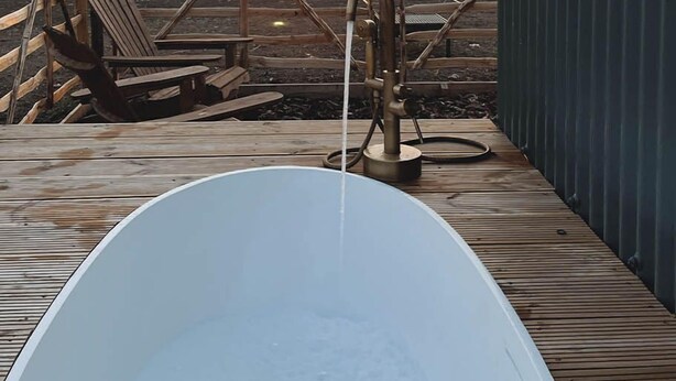 Eine Holzterrasse mit einer weißen Badewanne, einem Metallzaun und Blick auf den Sonnenuntergang über einem Feld.