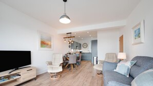 Un salon moderne avec un téléviseur à écran plat, un canapé gris et un coin repas avec une cuisine en arrière-plan.