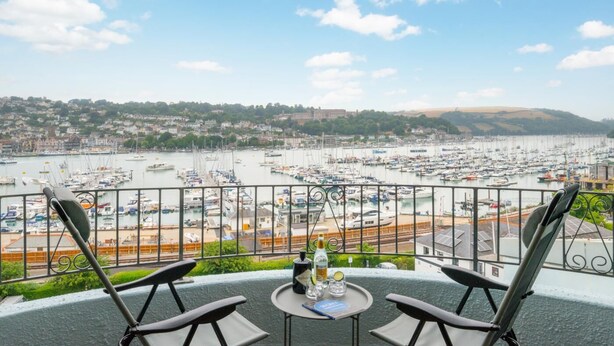 Un balcon avec deux chaises et une table, donnant sur une marina où sont amarrés de nombreux bateaux, avec une vue sur la ville en arrière-plan.