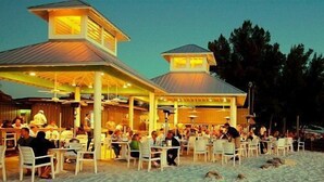 A beachside restaurant with outdoor seating and a covered area, overlooking the ocean.