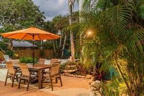 An outdoor patio with a wooden table and chairs, an orange umbrella, and tropical plants.