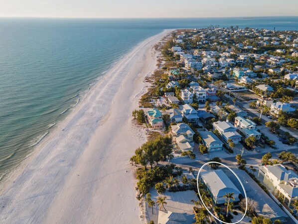 An aerial view of a coastal residential area with houses along the beach, a clear view of the ocean, and a distinct white building circled in the image.