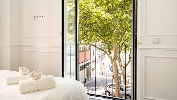 Ein Hotelzimmer mit großem Fenster, einem Bett mit weißen Laken und Blick auf einen Baum und die Straße.