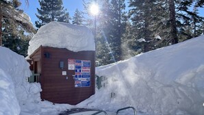 Une cabane en bois avec une piscine partiellement recouverte de neige, entourée de congères et bordée d’arbres.