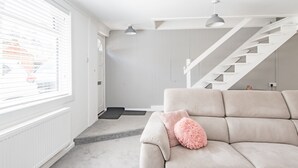 Een moderne woonkamer met een beige bank, een roze pluizig kussen, een trap en een raam met jaloezieën.
