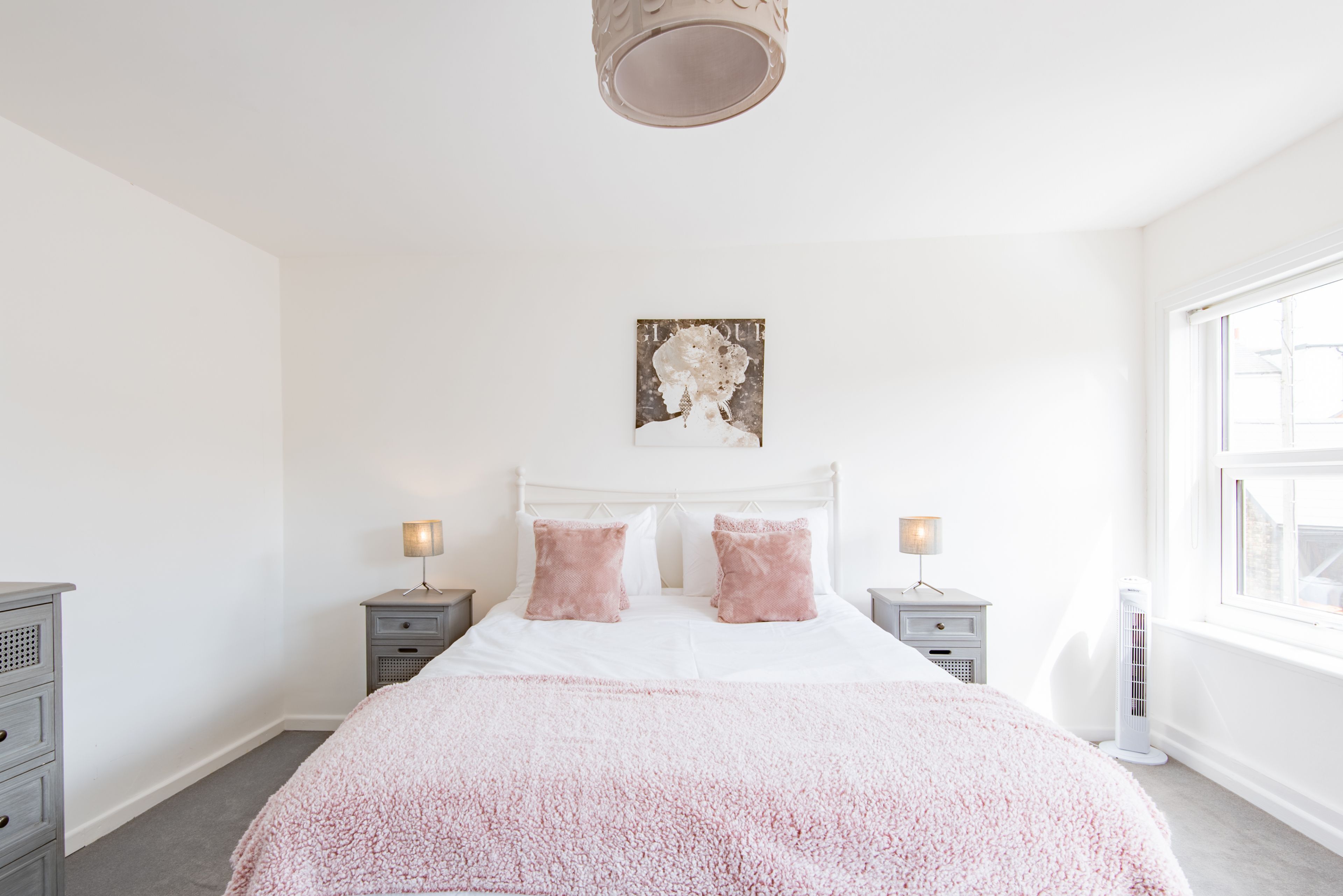 Une chambre à coucher avec un lit blanc, deux tables de chevet, une commode, une lampe fixée au mur et une fenêtre.