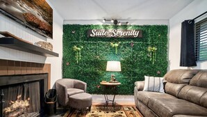 Ein Wohnzimmer mit Kamin, einer Couch, einem Sessel, einem Teppich sowie einer Wand mit Grünpflanzen und einem Schild mit der Aufschrift 'Suite Serenity'.
