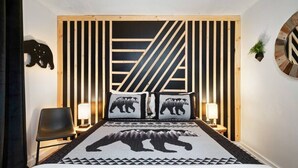 Ein Schlafzimmer mit einem Bett, zwei Nachttischlampen, einem Sessel, einer Wanduhr und einem Bären-Kissen.