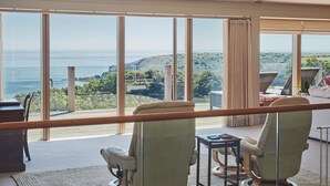 A spacious room with large windows offering a view of the sea and hills. There are two chairs and a small table.