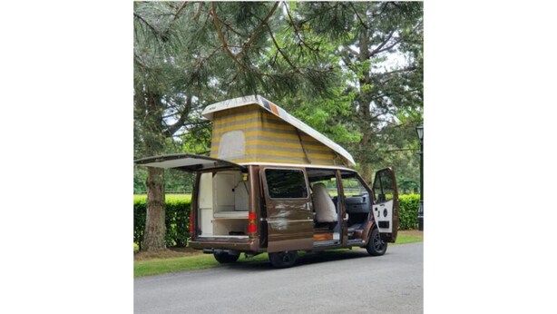 Una furgoneta marrón con un toldo grande y a rayas, y una puerta lateral abierta, estacionada en una calle con árboles al fondo.